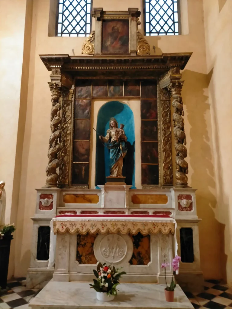 Vue d'un retable en marbre polychrome et bois doré à l'intérieur de la Cathédrale de la Nativité-de-Marie à Vence. Au centre, une statue de la Vierge Marie portant l'Enfant Jésus est placée dans une niche bleue. La statue est encadrée par deux colonnes torsadées richement sculptées et décorées de pampres de vigne. L'autel au premier plan est recouvert d'une nappe blanche brodée, devant laquelle se trouvent des bouquets de fleurs et une petite plante en pot. En arrière-plan, deux fenêtres à carreaux en losange éclairent la scène. @jeanpaul