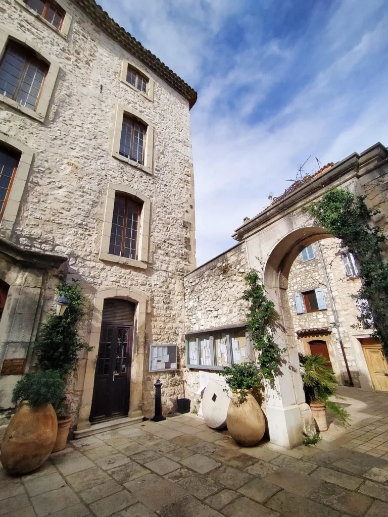 Vue prise depuis un porche voûté en pierre médiévale menant vers une place étroite du village de Tourrettes-sur-Loup. @jeanpaul 2025