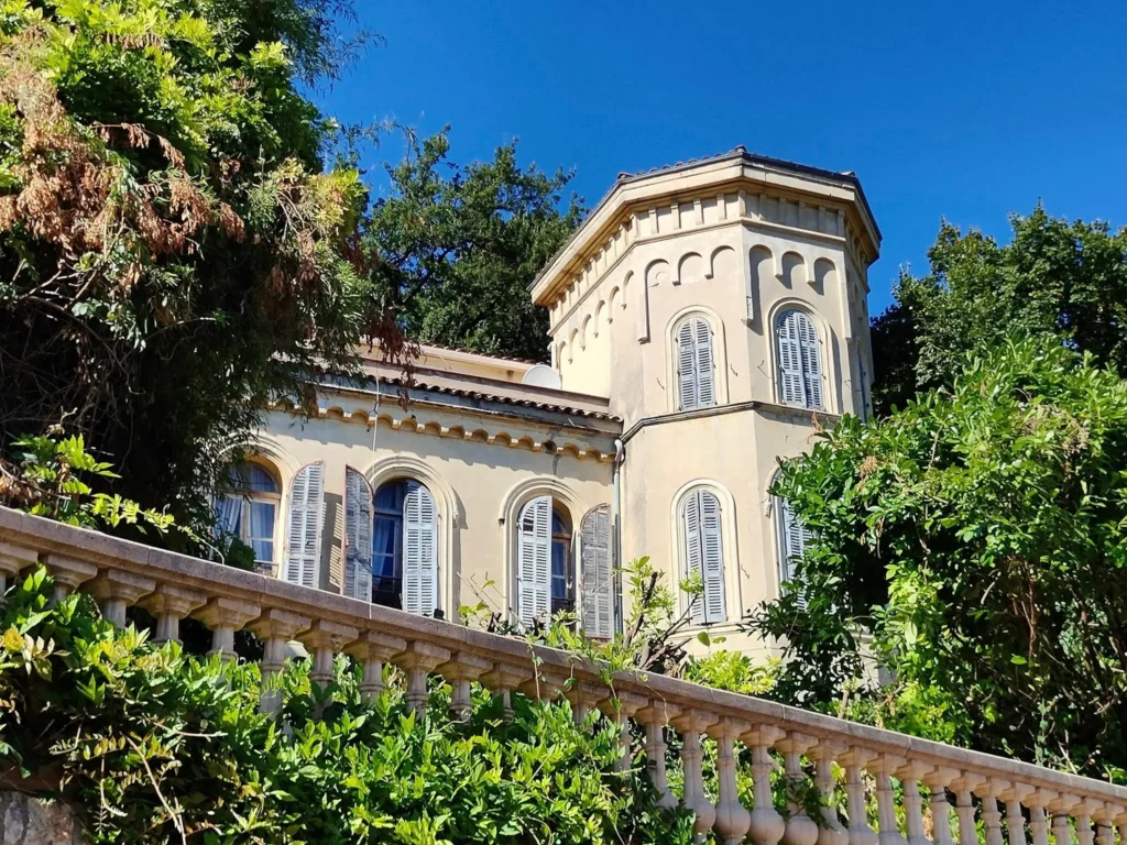 Façade d'une villa historique au Bar-sur-Loup avec une tour octogonale, des fenêtres à arcades et des volets bleus, entourée de végétation.