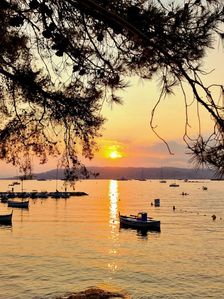 Une vue spectaculaire du littoral azuréen à l'heure dorée. Le soleil décline sur l'horizon, illuminant l'eau et découpant la silhouette d'un bateau au large de la baie.