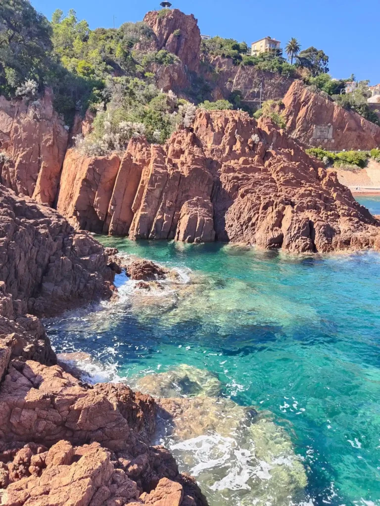Falaises rocheuses rouges accidentées du massif de l'Estérel s'élevant verticalement au-dessus d'une eau de mer turquoise et translucide dans une calanque sous un ciel bleu clair. @jeanpaul2025