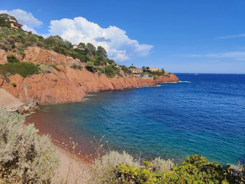 Petite baie sauvage aux eaux bleues bordée par une plage de sable ocre et les falaises rouges du massif de l'Estérel. @jeanpaul 2025