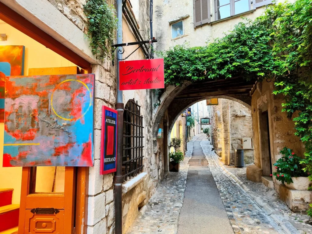 Vue prise depuis l'entrée d'une ruelle médiévale étroite et pavée à Saint-Paul-de-Vence. Les façades en pierre dorée des bâtiments sont ornées de plantes grimpantes. De chaque côté, des enseignes en fer forgé signalent des galeries d'art et des ateliers d'artistes. La rue monte doucement vers une arche en arrière-plan sous un ciel bleu clair. @jeanpaul 2025
