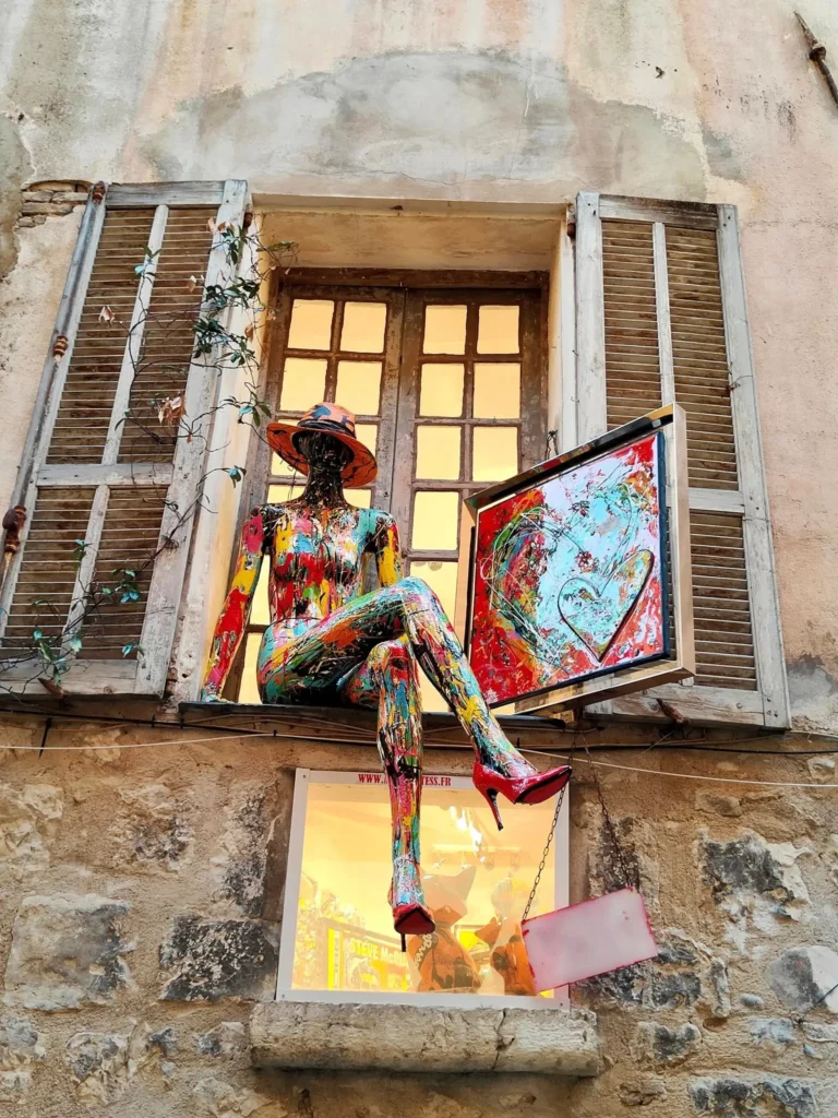 Un mannequin de femme assis, peint de manière artistique avec des éclaboussures de peinture multicolores et portant un chapeau orange et des talons aiguilles rouges, est perché sur le rebord de la fenêtre en bois d'un bâtiment en pierre à Saint-Paul-de-Vence. Le rebord de la fenêtre est éclairé de l'intérieur, et le mannequin tient un tableau encadré d'un cœur rouge stylisé. Les volets en bois anciens encadrent la fenêtre. Le mur est en pierre naturelle. @jeanpaul2025