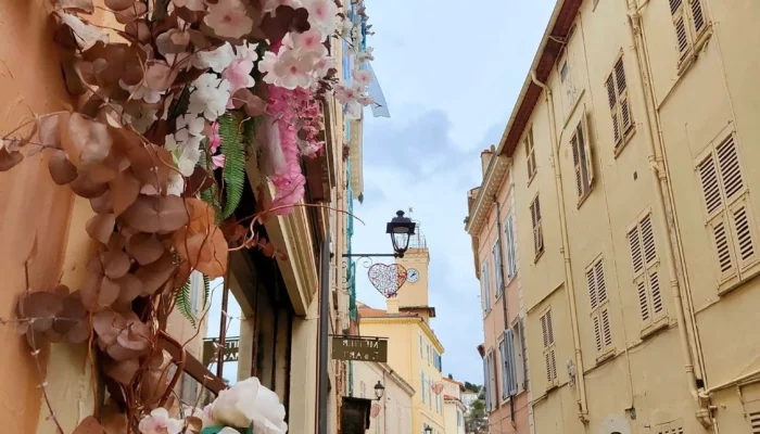 Vue en perspective d'une ruelle étroite et pavée du Vieux-Cannet, la rue Saint-Sauveur, bordée de bâtiments provençaux aux façades ocre et jaune. Au premier plan à gauche, une composition florale dense de fleurs séchées marron et de fleurs artificielles roses et blanches (pivoines et hortensias) encadre une vitrine. Au loin, dans l'axe de la rue, se dresse le clocher jaune emblématique de la Chapelle Saint-Sauveur sous un ciel couvert, avec un cœur décoratif visible près du clocher. @jeanpaul 2025