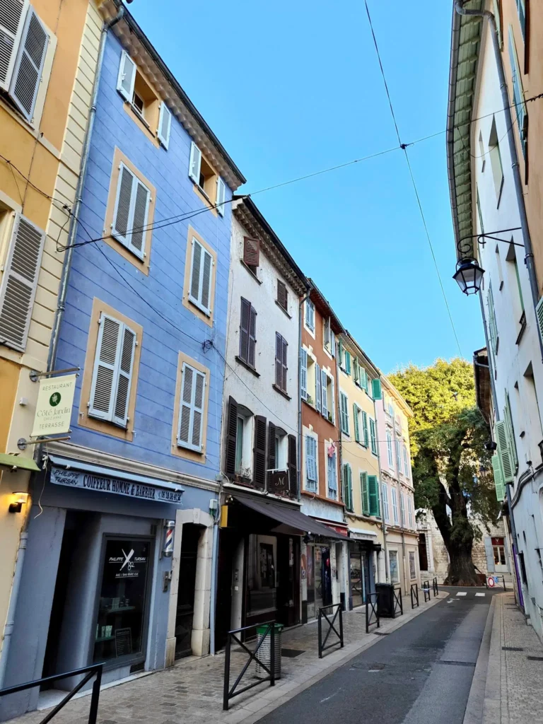 ue typique du centre historique de Vence avec ses façades colorées (ocre, bleu, jaune), ses commerces locaux comme le coiffeur Philippe Tursan et le restaurant Côté Jardin, sous un ciel bleu azur. Au bout de la ruelle, on aperçoit le célèbre frêne de la place. @jeanpaul