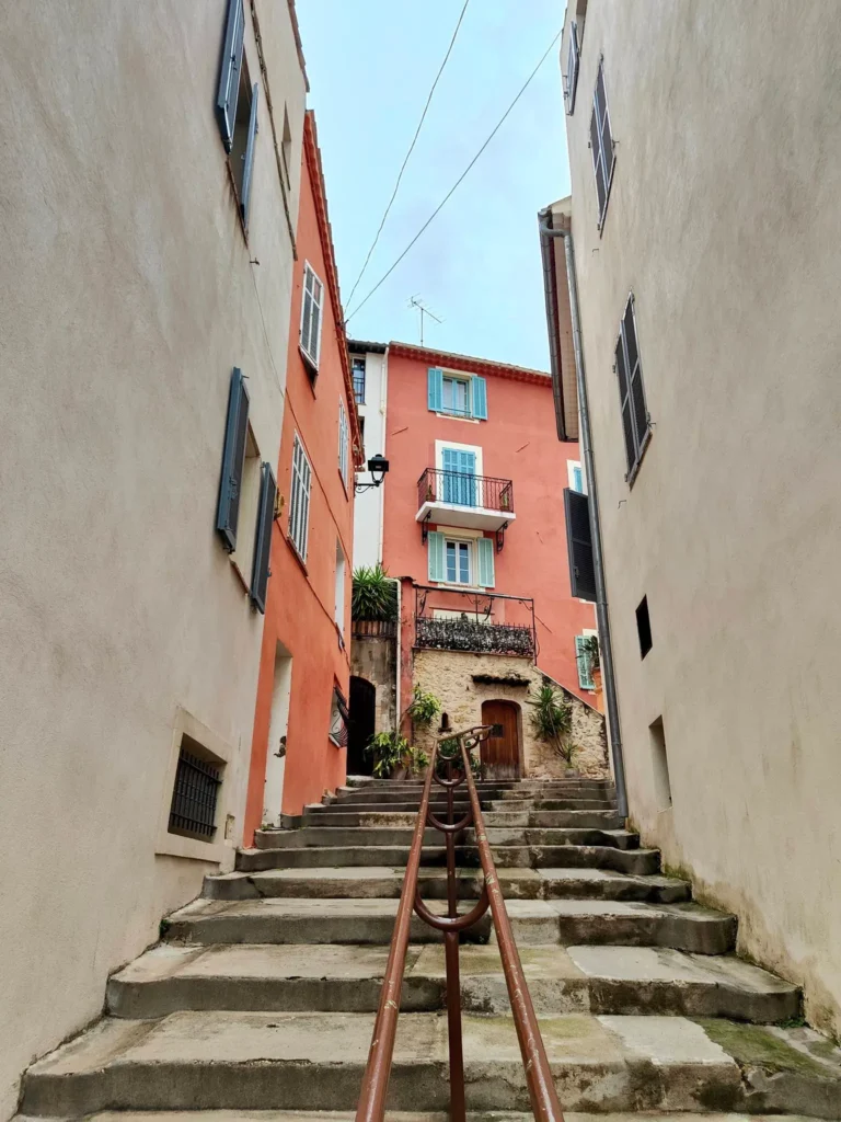 ue en contre-plongée des escaliers en pierre du Vieux-Cannet, avec une rampe centrale en fer forgé. La montée est encadrée par des façades provençales aux couleurs ocre, rose et blanc, dotées de volets bleus et gris, sous un ciel clair. @jeanpaul 2025