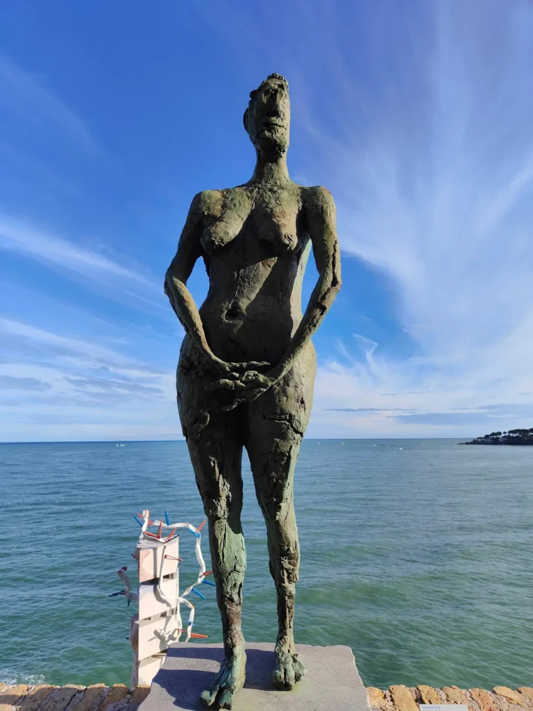 Vue depuis la terrasse du Musée Picasso à Antibes. Au premier plan, une imposante statue de femme nue en bronze, patinée par le temps, est debout face à la mer, les mains jointes. En arrière-plan, la mer Méditerranée s'étend à l'horizon sous un ciel bleu vif avec des nuages légers. Les remparts en pierre du Château Grimaldi encadrent la scène. @jeanpaul 2025