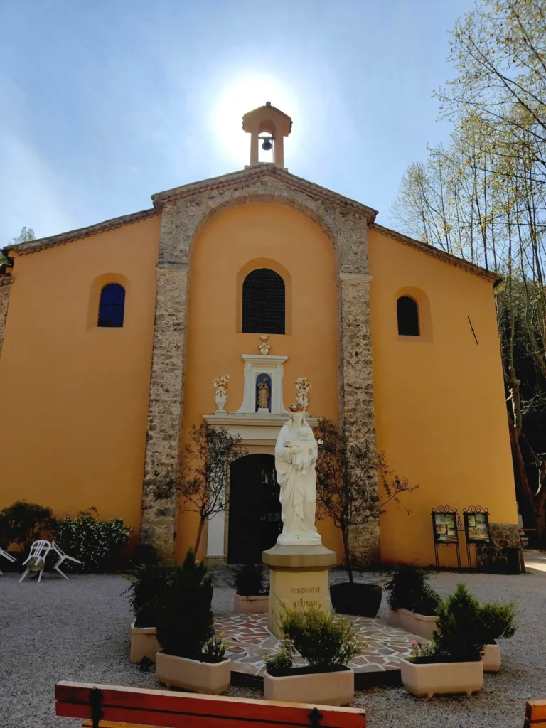 Vue de face de la façade principale du Sanctuaire Notre-Dame de Vaucluse à Auribeau-sur-Siagne, sous un ciel bleu clair avec le soleil brillant directement au-dessus du clocher-mur central. @jeanpaul 2025
