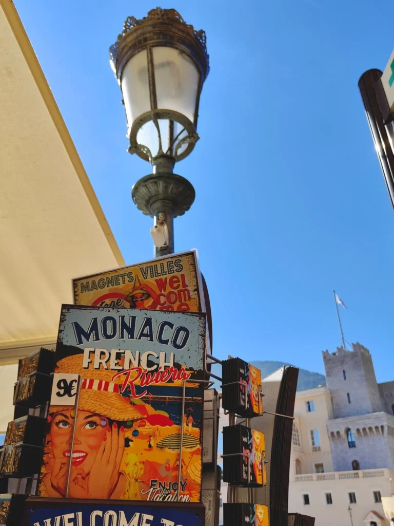 Une vue verticale d'un présentoir de souvenirs accroché à un lampadaire à gaz orné à Monaco. L'affiche en métal principale montre une femme de style vintage avec le texte "MONACO" et "FRENCH RIVIERA", ainsi que "Enjoy Your Vacation". En dessous, on trouve des cartes postales plus petites. En arrière-plan, une tour de château médiéval en pierre avec un drapeau se dresse contre un ciel bleu clair, à côté d'un bâtiment avec une façade beige et un auvent. @jeanpaul 2026