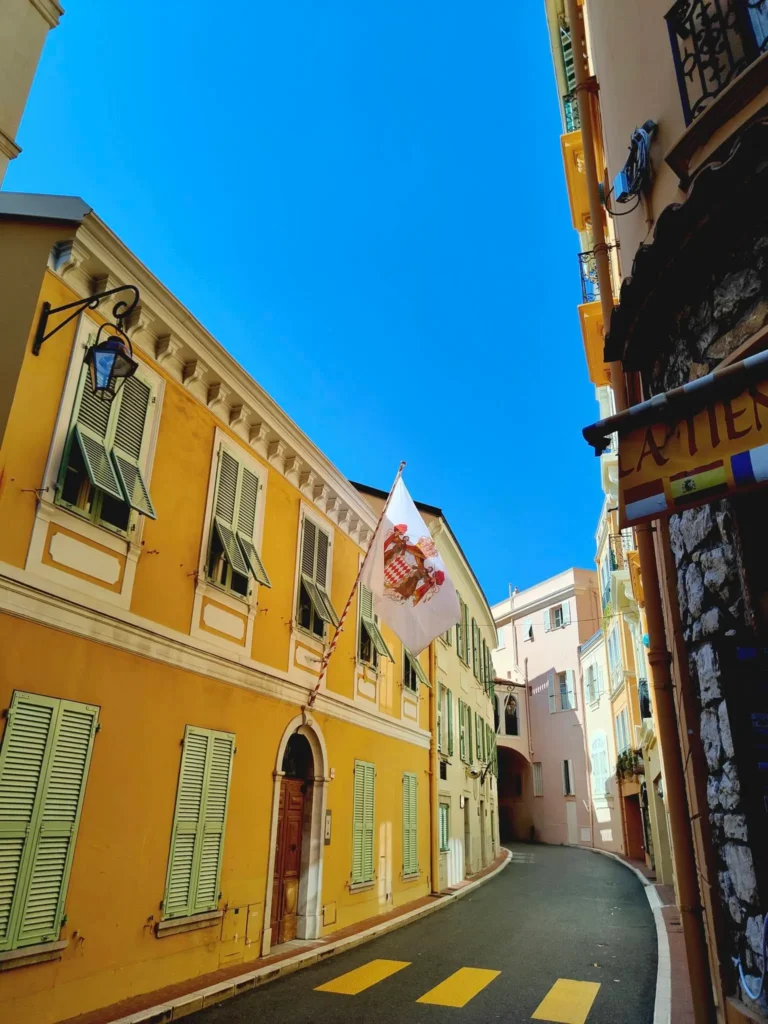 Perspective montante sur une ruelle pavée et sinueuse de Monaco, bordée de façades colorées en ocre jaune et rose pastel. Sur la gauche, un bâtiment jaune vif arbore des fenêtres ornées de volets persiennes vert amande entrouverts. Un grand drapeau blanc aux armoiries rouges et dorées de Monaco est suspendu au-dessus d'une porte d'entrée en bois. Au premier plan à droite, un mur en pierres apparentes porte une enseigne partielle. La rue est délimitée par un passage piéton jaune au sol, le tout sous un ciel bleu azur intense et dégagé. @jeanpaul 2025