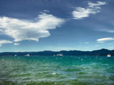 Une vue sur les eaux calmes et azur de la mer de Saint-Tropez près de la plage des Salins. Le soleil fait scintiller la surface de la mer Méditerranée, bordée au loin par un littoral sauvage composé de collines verdoyantes, de pins parasols et de maquis provençal. Le ciel est entièrement bleu et dégagé, offrant une atmosphère de farniente typique de la Riviera.