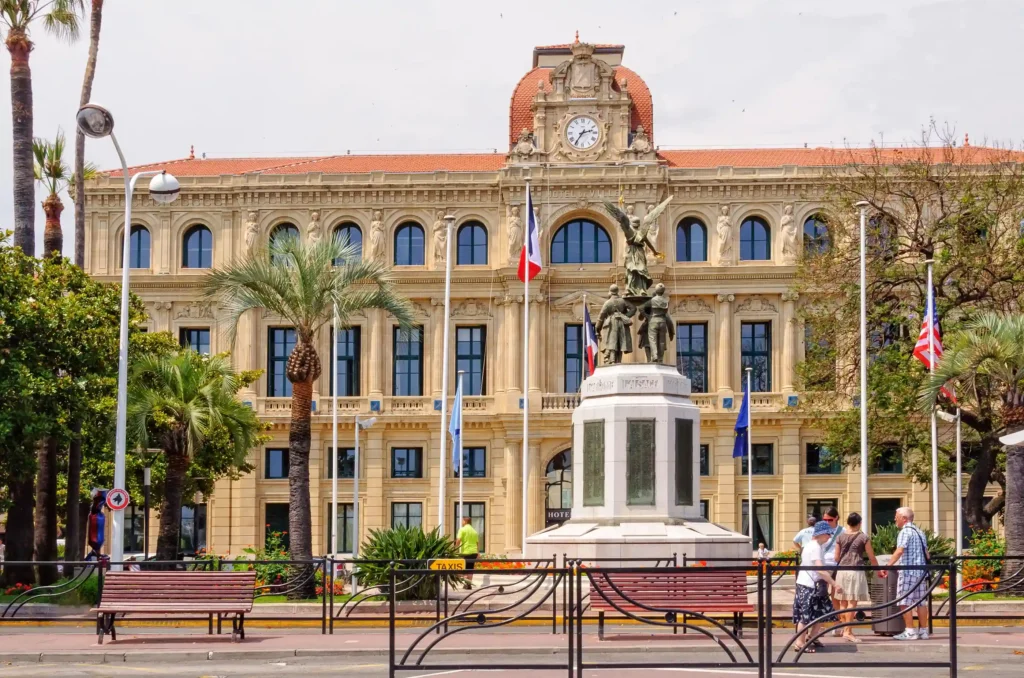Vue d'angle de l'Hôtel de Ville de Cannes, un imposant bâtiment néo-classique en pierre calcaire dorée. La photo montre sa longue façade latérale rythmée par des arcades en plein cintre et des pilastres. Son toit de tuiles plates orange et le clocher central avec horloge sont bien visibles sous un ciel clair. Un grand affichage d'art noir et blanc orne l'angle.