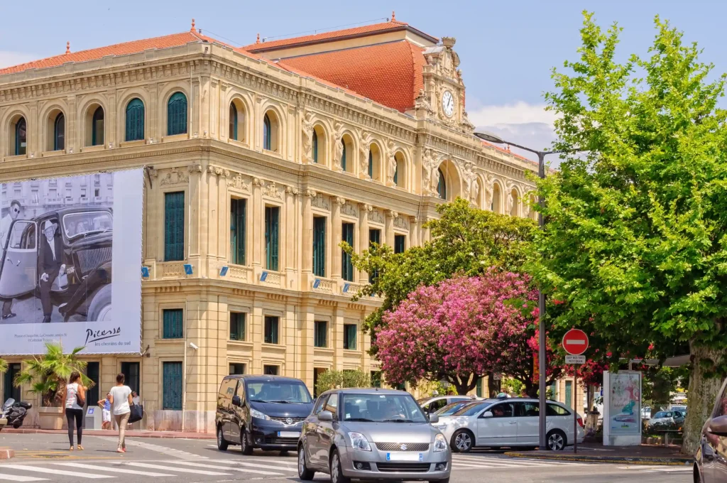 Vue du magnifique bâtiment historique de l'Hôtel de Ville de Cannes, à l'architecture néo-classique dorée et aux toits de tuiles oranges, sous un ciel partiellement nuageux. À l'angle, une immense affiche d'art en noir et blanc représente Picasso dans une voiture ancienne, avec l'inscription "Picasso" et "les chemins du sud". Le bâtiment fait face à une rue pavée et à un carrefour où des voitures circulent, des arbres verts et un arbre à fleurs roses sont visibles. Deux piétons marchent sur le trottoir à gauche. 