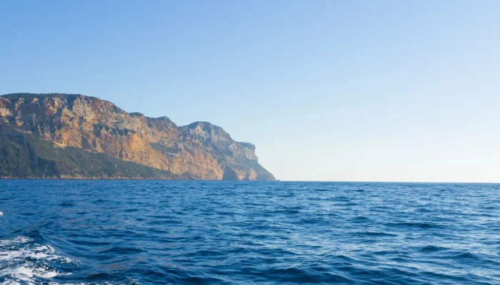 Spectacular panoramic view of Cap Canaille. The ochre limestone cliffs plunge vertically into the deep blue waters of the Mediterranean Sea. In the background, the bay of La Ciotat stretches out under a vast sunny sky, bordered by the coastline of the French Riviera.