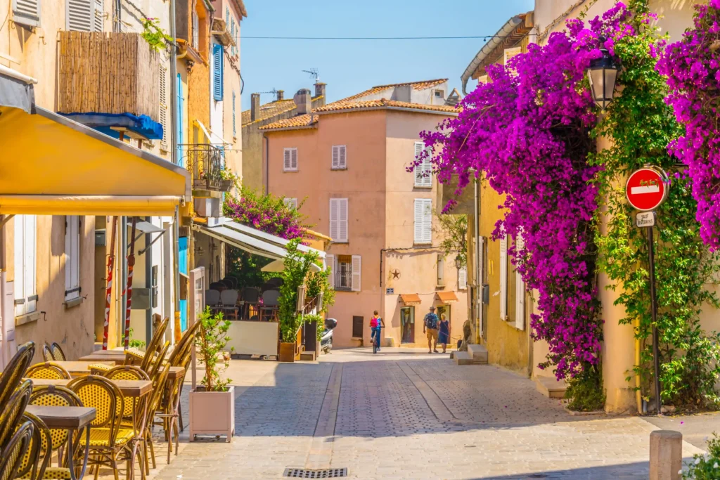 Vue d'une ruelle pavée ensoleillée dans la vieille ville de Saint-Tropez. À gauche, la terrasse d'un restaurant typique avec ses tables et chaises en rotin alignées sous un store jaune. À droite, une magnifique cascade de bougainvilliers d'un violet éclatant grimpe le long d'une façade ocre, à côté d'un panneau de signalisation. En arrière-plan, quelques passants marchent sous un grand ciel bleu.
