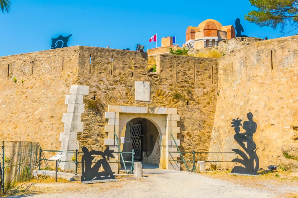 Photographie de l'entrée fortifiée de la Citadelle de Saint-Tropez. On y voit une porte d'entrée en pierre de taille blanche en arc de cercle avec un pont-levis en bois. De hauts murs défensifs en pierre ocre entourent l'entrée, surmontés d'un cadran solaire et du drapeau français. À droite, un ancien canon en bronze est exposé sur son affût en bois, et à gauche, trois oliviers sont plantés dans des pots en terre cuite.