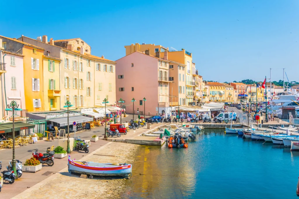 ue panoramique sur le Vieux Port de Saint-Tropez animé. Au premier plan, l'eau calme de la marina accueille des pointus traditionnels en bois coloré amarrés aux côtés de grands yachts de luxe modernes. En arrière-plan, les célèbres façades de maisons de couleur pastel, ocre et rose s'alignent le long des quais, animées par les terrasses de cafés. 