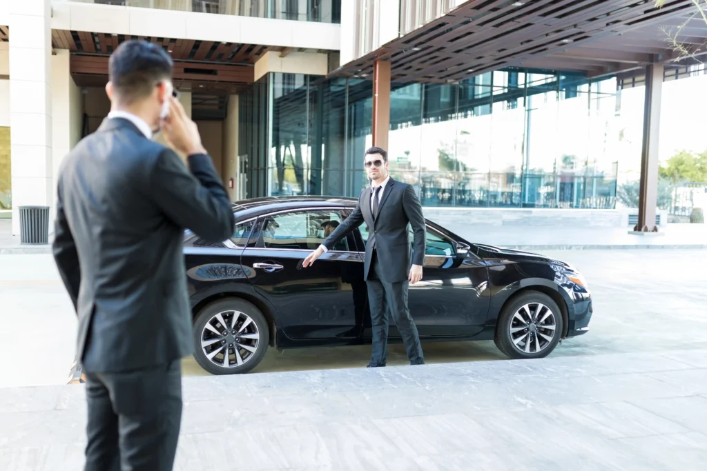 Agent de protection rapprochée effectuant l'ouverture de porte pour un client VIP. Chauffeur de sécurité en costume et kit oreillette, devant une berline noire de fonction. Protocole d'embarquement sécurisé.
