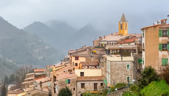 Village perché de Sainte-Agnès avec ses maisons en pierre, son clocher jaune et les montagnes environnantes sous un ciel brumeux.