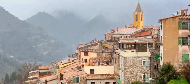 Village perché de Sainte-Agnès avec ses maisons en pierre, son clocher jaune et les montagnes environnantes sous un ciel brumeux.