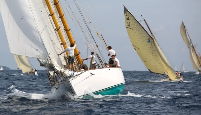 Vue rapprochée d’un grand voilier classique blanc aux mâtures en bois verni fendant les vagues en pleine mer comme lors des voiles de saint tropez. L’équipage vêtu de t-shirts blancs s’active sur le pont et au pied du mât pendant la manœuvre sous de grandes voiles blanches déployées. À l’arrière-plan, deux autres voiliers d’époque aux voiles jaunies inclinées par le vent naviguent sur une mer bleue agitation moyenne avec de l’écume blanche sous un ciel lumineux.