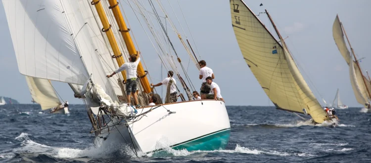 Vue rapprochée d’un grand voilier classique blanc aux mâtures en bois verni fendant les vagues en pleine mer comme lors des voiles de saint tropez. L’équipage vêtu de t-shirts blancs s’active sur le pont et au pied du mât pendant la manœuvre sous de grandes voiles blanches déployées. À l’arrière-plan, deux autres voiliers d’époque aux voiles jaunies inclinées par le vent naviguent sur une mer bleue agitation moyenne avec de l’écume blanche sous un ciel lumineux.