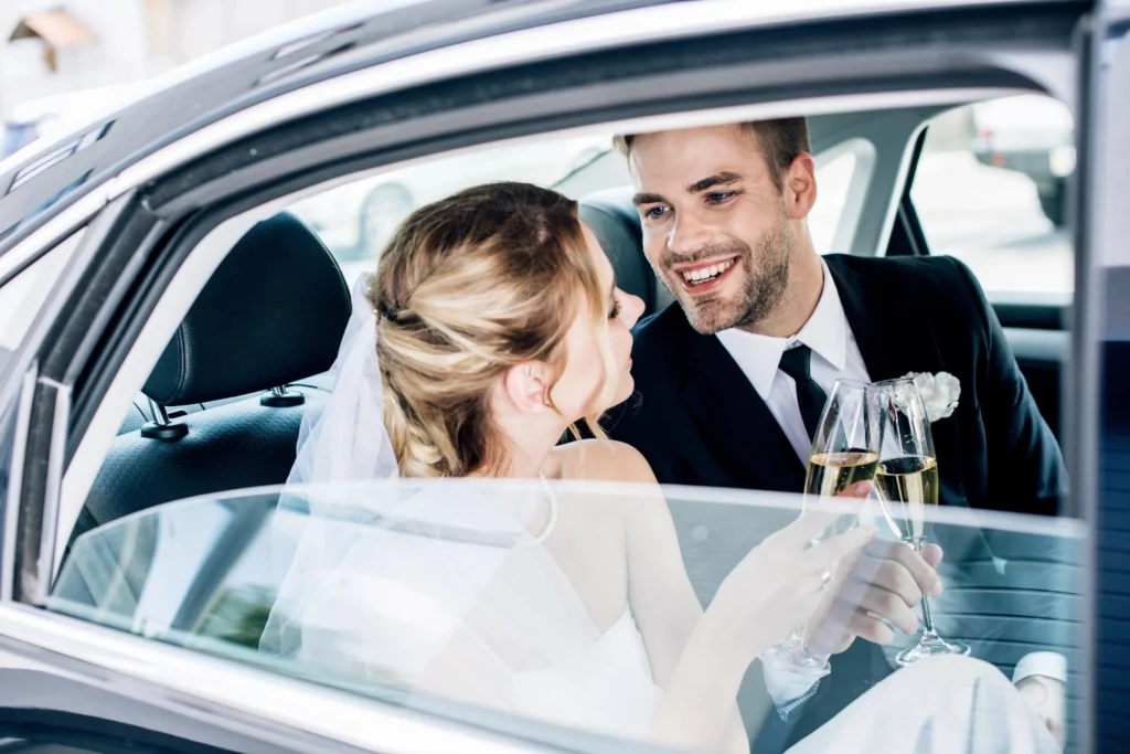 Jeunes mariés souriants trinquant avec des flûtes de champagne à l'arrière d'une voiture de luxe avec chauffeur privé pour mariage.
