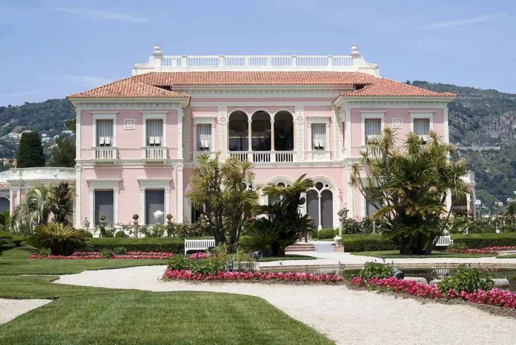 Vue de face de la façade symétrique rose et blanche de style Renaissance italienne au cours d'une visite de la Villa Ephrussi de Rothschild. Le palais rose se dresse derrière un jardin à la française verdoyant, orné de parterres de fleurs roses, d'allées en gravier blanc, de palmiers exotiques et de buissons d'ornement. L'étage supérieur présente une loggia ouverte avec des arches triples et des balustrades blanches sous un toit de tuiles romaines en terre cuite. En arrière-plan, les collines rocheuses de la Côte d'Azur se dessinent sous un ciel bleu clair.