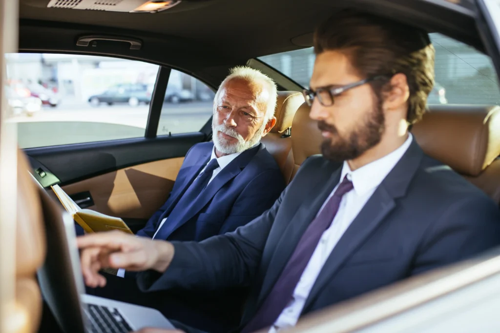 Deux hommes d'affaires en costume travaillant à l'arrière d'une berline de luxe avec sièges en cuir beige. L'un utilise un ordinateur portable tandis que l'autre consulte un carnet de notes.La mobilité executive sans compromis. EDEN CAB assure vos transferts entre l'aéroport de Nice et vos centres de congrès à Cannes, en vous garantissant discrétion et confort pour vos échanges professionnels.