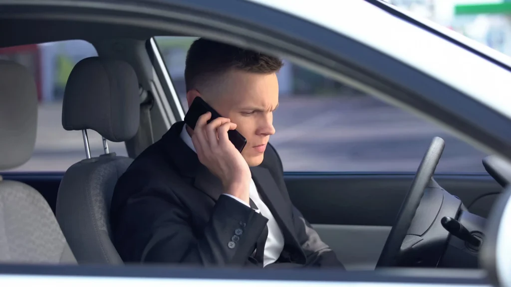 Chauffeur de sécurité en costume noir à l'intérieur d'un véhicule, communiquant avec attention par téléphone mobile. Image illustrant la coordination logistique et la réactivité du service de transport VIP.