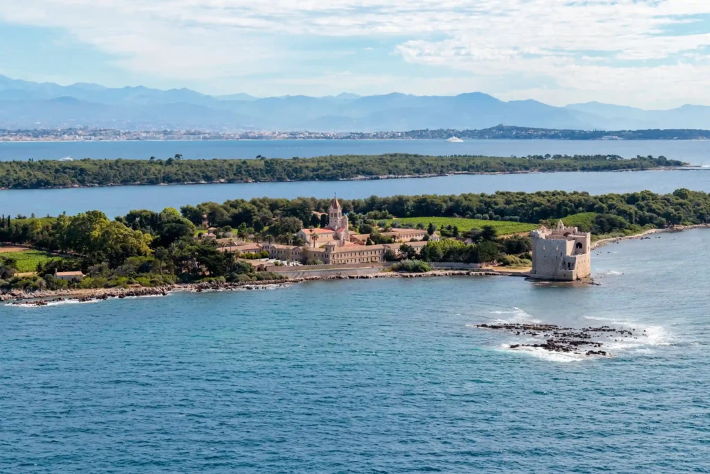 Vue aérienne panoramique de l'île Saint-Honorat, au large de Cannes. L'île, recouverte d'une végétation dense et de vignobles, est entourée d'une mer turquoise et transparente. Au premier plan à droite, le monastère fortifié médiéval en pierre se dresse sur la côte rocheuse. Plus loin, au centre, se trouvent l'abbaye moderne et son clocher. En arrière-plan, la côte cannoise et les montagnes de l'Esterel se dessinent sous un ciel clair.