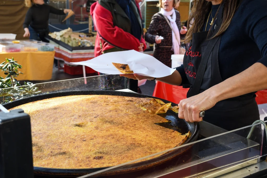 Photographie en gros plan d'une marchande préparant de la socca sur un marché à Cannes. Elle porte un tablier noir avec "Socca du Coeur" brodé en rouge. Sa main droite, munie d'un couteau, découpe une part d'une grande et fine crêpe de farine de pois chiche, bien dorée, qui cuit dans une poêle ronde en métal de grand diamètre. La part découpée est posée sur un papier blanc, prête à être servie. Le stand est équipé d'une plaque de protection en verre. En arrière-plan, d'autres stands de marché avec des nappes jaunes et rouges sont visibles, ainsi que quelques clients. L'éclairage est naturel.