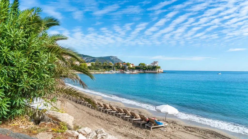 Vue de la plage de la Baie des Fourmis à Beaulieu-sur-Mer avec chaises longues, parasol et la Villa Kérylos en arrière-plan sous un ciel nuageux.