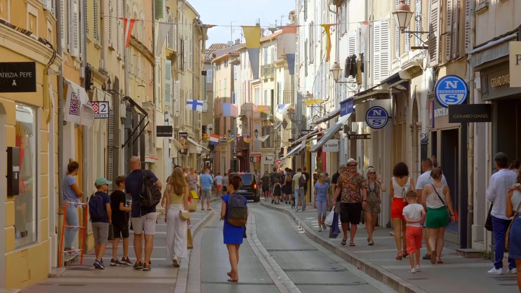 Vue en perspective d'une ruelle pavée et ombragée de la vieille ville de Saint-Tropez. Des passants et des visiteurs flânent le long des façades anciennes aux couleurs chaudes, ocre et pastel, ornées de volets en bois et de balcons fleuris de bougainvilliers.