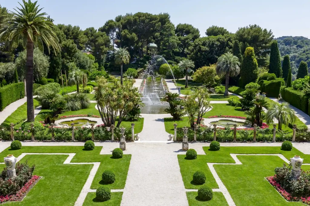 Vue surélevée et panoramique du jardin à la française conçu en forme de pont de navire, prise lors d'une visite de la Villa Ephrussi de Rothschild. Une large allée centrale en gravier blanc traverse des pelouses d'un vert éclatant, ornées de buis taillés en boules symétriques. Au centre, un long canal d'eau rectiligne présente de grands jets d'eau en cascade. Le canal mène à un escalier et, en arrière-plan sur les hauteurs, au Temple de l'Amour entouré de grands arbres et d'une forêt de pins. Le jardin est parsemé de palmiers exotiques, de parterres de fleurs et de balustrades en pierre décorées de vases en porcelaine.