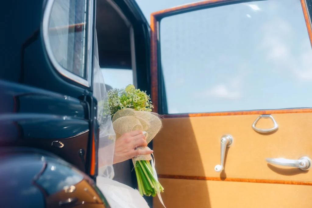 Main de la mariée tenant son bouquet de fleurs depuis la portière ouverte d'une voiture de collection, parfaite pour une décoration voiture des mariés élégante.