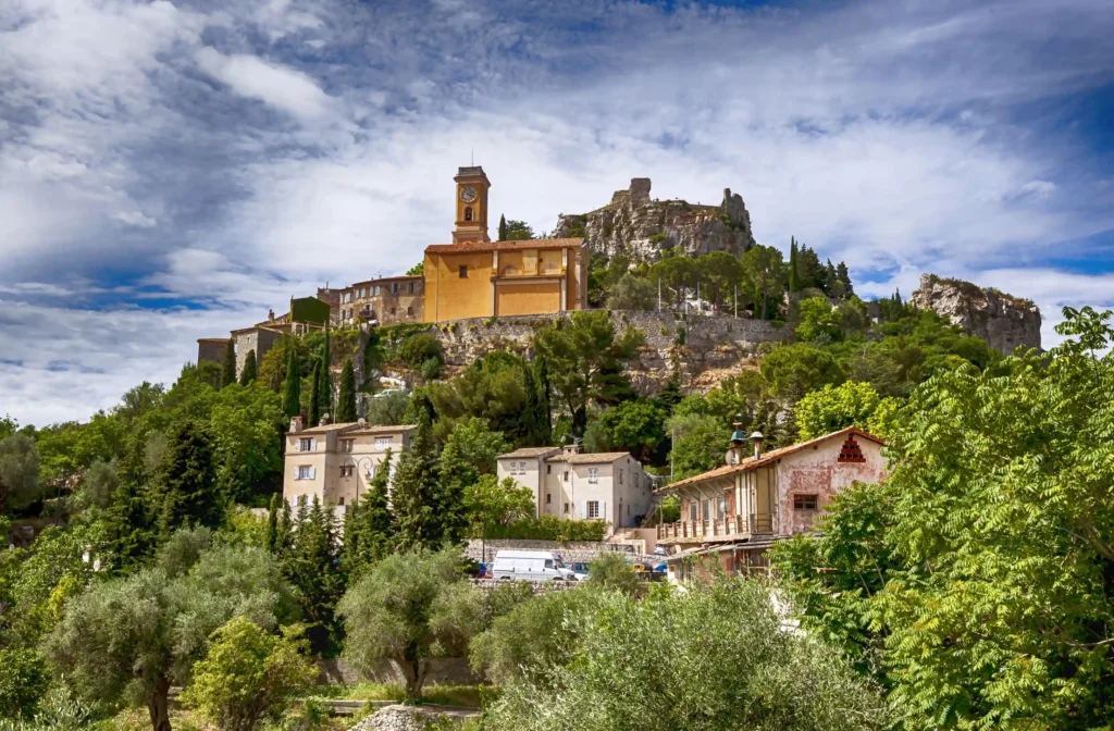 Vue panoramique en contre-plongée du village médiéval perché d'Èze. Au sommet de la colline rocheuse se détache l'église Notre-Dame-de-l'Assomption avec son clocher ocre jaune et son horloge, surmontée plus haut par les ruines du château fort. Les flancs de la colline sont recouverts d'une végétation dense d'arbres, de pins et de cyprès, au milieu desquels s'intègrent des maisons provençales en pierre. Le ciel bleu est partiellement couvert de nuages blancs moutonneux.