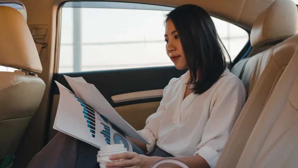 Une femme d'affaires travaillant sur des documents financiers à l'arrière d'un véhicule de luxe conduit par un chauffeur privé VIP.