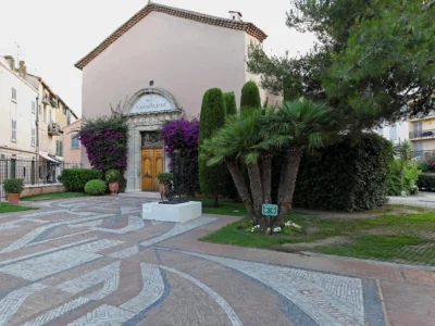 Vue de la façade extérieure du musée de l'annonciade saint tropez. L'ancienne chapelle aux murs beige rosé présente une entrée en arc de pierre surmontée de l'inscription du musée, avec une double porte en bois. Le porche est encadré par de magnifiques bougainvilliers violets en fleurs et de hauts cyprès. Au premier plan, un grand parvis pavé de motifs géométriques en mosaïque est bordé de palmiers et de végétation méditerranéenne.