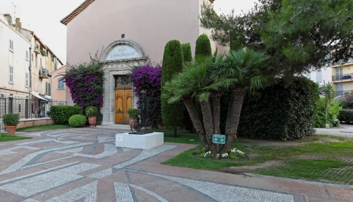 Vue de la façade extérieure du musée de l'annonciade saint tropez. L'ancienne chapelle aux murs beige rosé présente une entrée en arc de pierre surmontée de l'inscription du musée, avec une double porte en bois. Le porche est encadré par de magnifiques bougainvilliers violets en fleurs et de hauts cyprès. Au premier plan, un grand parvis pavé de motifs géométriques en mosaïque est bordé de palmiers et de végétation méditerranéenne.