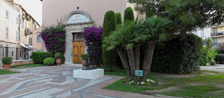 Vue de la façade extérieure du musée de l'annonciade saint tropez. L'ancienne chapelle aux murs beige rosé présente une entrée en arc de pierre surmontée de l'inscription du musée, avec une double porte en bois. Le porche est encadré par de magnifiques bougainvilliers violets en fleurs et de hauts cyprès. Au premier plan, un grand parvis pavé de motifs géométriques en mosaïque est bordé de palmiers et de végétation méditerranéenne.