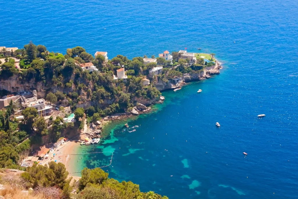 Vue aérienne plongeante et panoramique sur la rade de Villefranche-sur-Mer. Au premier plan, des eaux cristallines en dégradé de turquoise et de bleu profond accueillent de nombreux voiliers au mouillage. À droite, les façades colorées aux tons ocre, jaune et orange de la vieille ville bordent le littoral, dominées par des collines verdoyantes sous un grand ciel bleu.