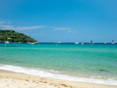 Vue sur les eaux turquoise et cristallines de la plage de Pampelonne à Ramatuelle. Au premier plan, de légères vagues d'écume blanche viennent lécher le sable fin et doré. Au large, sous un ciel bleu azur et dégagé, plusieurs yachts et bateaux de plaisance sont ancrés dans la baie. Sur la gauche, une colline verdoyante recouverte de pins et de végétation borde le littoral.