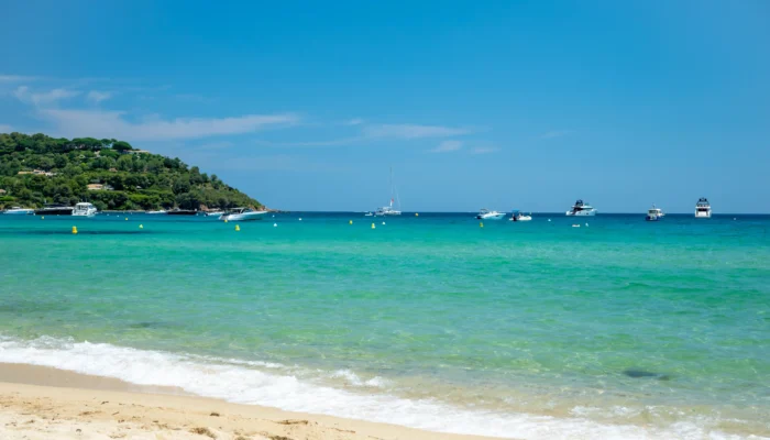 Vue sur les eaux turquoise et cristallines de la plage de Pampelonne à Ramatuelle. Au premier plan, de légères vagues d'écume blanche viennent lécher le sable fin et doré. Au large, sous un ciel bleu azur et dégagé, plusieurs yachts et bateaux de plaisance sont ancrés dans la baie. Sur la gauche, une colline verdoyante recouverte de pins et de végétation borde le littoral.