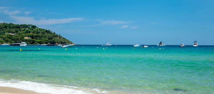 Vue sur les eaux turquoise et cristallines de la plage de Pampelonne à Ramatuelle. Au premier plan, de légères vagues d'écume blanche viennent lécher le sable fin et doré. Au large, sous un ciel bleu azur et dégagé, plusieurs yachts et bateaux de plaisance sont ancrés dans la baie. Sur la gauche, une colline verdoyante recouverte de pins et de végétation borde le littoral.