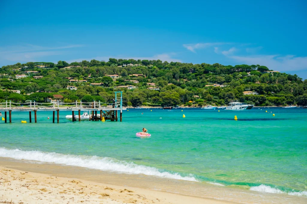 Vue de la plage de Pampelonne à Ramatuelle avec une eau translucide dégradée du vert émeraude au bleu turquoise. Au premier plan, une petite vague de mer vient lécher le sable doré. Sur la gauche, un long ponton en bois sur pilotis s'avance dans l'eau, à côté d'une petite embarcation. Au centre, une personne se détend sur une bouée gonflable rose. En arrière-plan, une colline verdoyante couverte de pins et parsemée de villas surplombe la baie sous un ciel azur.