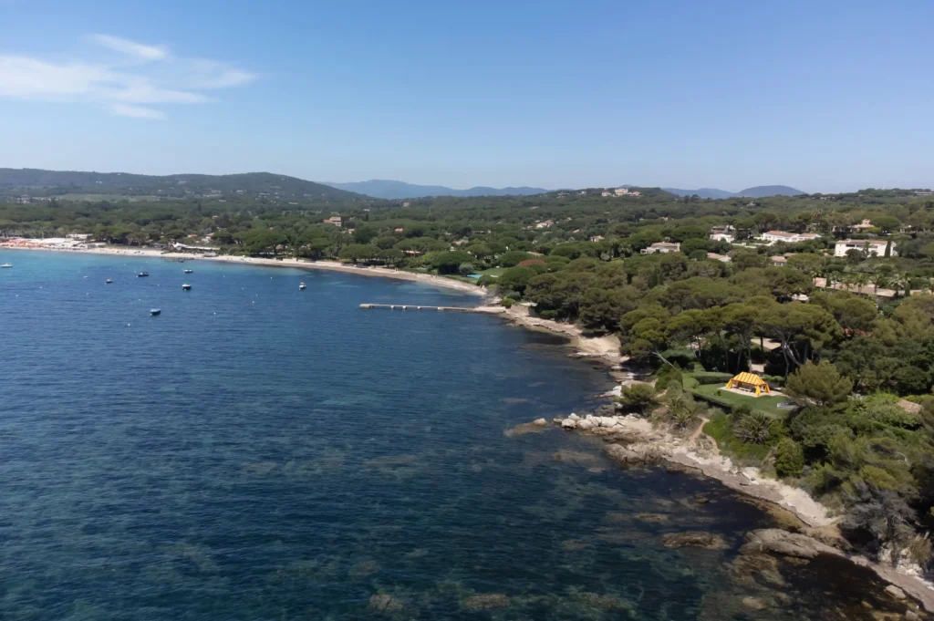 Vue aérienne en plongée de la baie de Pampelonne et de sa côte rocheuse et sauvage. L'eau transparente laisse deviner des fonds marins sombres et rocheux au premier plan, avant de s'étendre en un bleu profond parsemé de quelques embarcations au mouillage. Le littoral boisé est recouvert d'une immense pinède verdoyante où se nichent de somptueuses propriétés cachées par la végétation. Au loin, les reliefs des collines de Ramatuelle se dessinent sous un ciel bleu clair et dégagé.