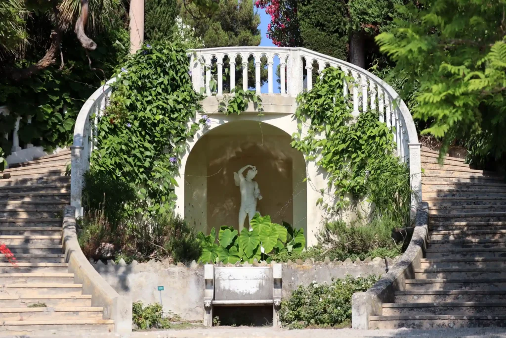 Vue de face d'une structure architecturale en pierre blanche dans les jardins, immortalisée lors d'une visite de la Villa Ephrussi de Rothschild. Au centre, une niche en arche abrite une statue en marbre blanc représentant un personnage mythologique debout. Cette alcôve est entourée de larges feuilles vertes au sol et encadrée par deux escaliers en pierre courbés qui montent de chaque côté vers un balcon en terrasse supérieure doté d'une balustrade blanche. Des plantes grimpantes et du lierre recouvrent en partie les flancs de la structure. Au premier plan, un banc en pierre patinée fait face à l'ensemble sous une lumière ensoleillée.