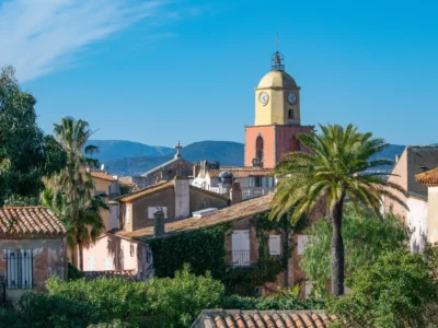Vue sur le célèbre clocher de style baroque de l'Église Notre-Dame-de-l’Assomption à Saint-Tropez. La tour du clocher affiche ses couleurs emblématiques jaune ocre et terre de Sienne, surmontée d'une horloge et d'un campanile en fer forgé. La structure se détache sur un ciel azur lumineux, entourée par les toits de tuiles provençales du village.